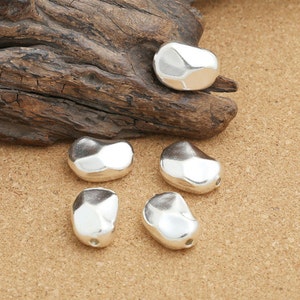 May include: Five silver-colored, irregularly shaped beads, resembling pebbles, are arranged on a brown, textured surface.