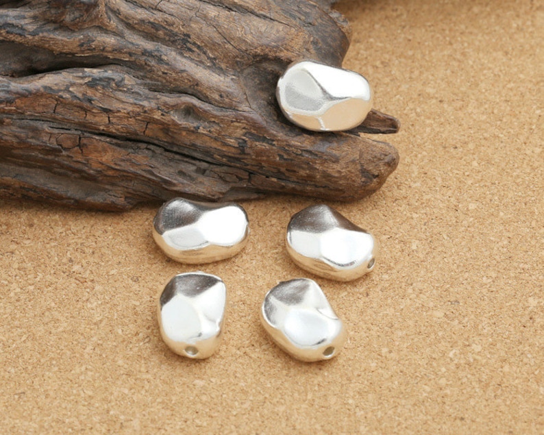 May include: Five silver-colored, irregularly shaped beads, resembling pebbles, are arranged on a brown, textured surface.