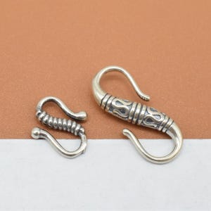 Könnte beinhalten: Zwei silberfarbene Verschlüsse zum Schmuckbasteln. Ein Verschluss ist schlicht, der andere hat ein dekoratives Design.