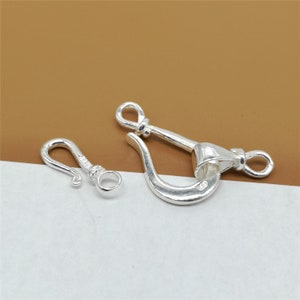 May include: Two silver-colored jewelry clasps. One is a small hook clasp, and the other is a larger shackle clasp. Both clasps have a shiny, metallic finish and are designed for jewelry making or repair. The clasps are displayed on a white and brown background.