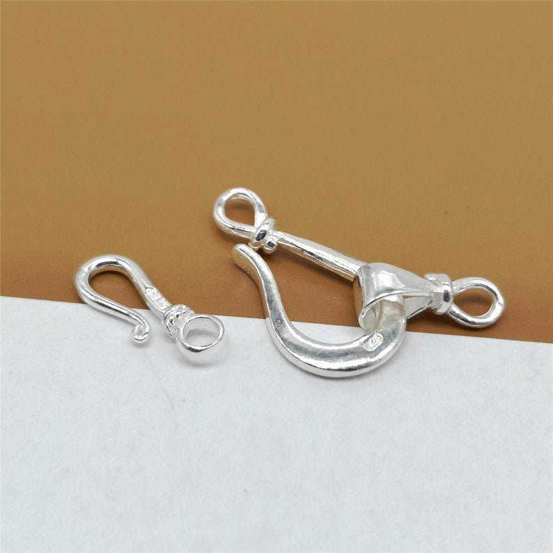 May include: Two silver-colored jewelry clasps. One is a small hook clasp, and the other is a larger shackle clasp. Both clasps have a shiny, metallic finish and are designed for jewelry making or repair. The clasps are displayed on a white and brown background.