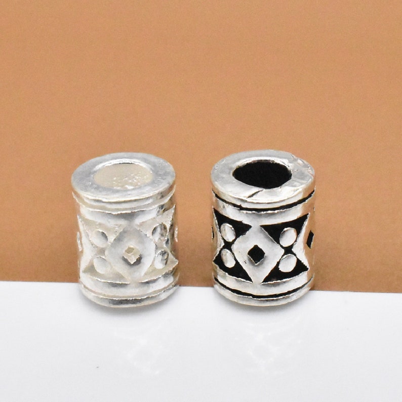 May include: Two silver hair beads with a geometric design. One bead is silver with a black design, and the other is silver with a silver design.