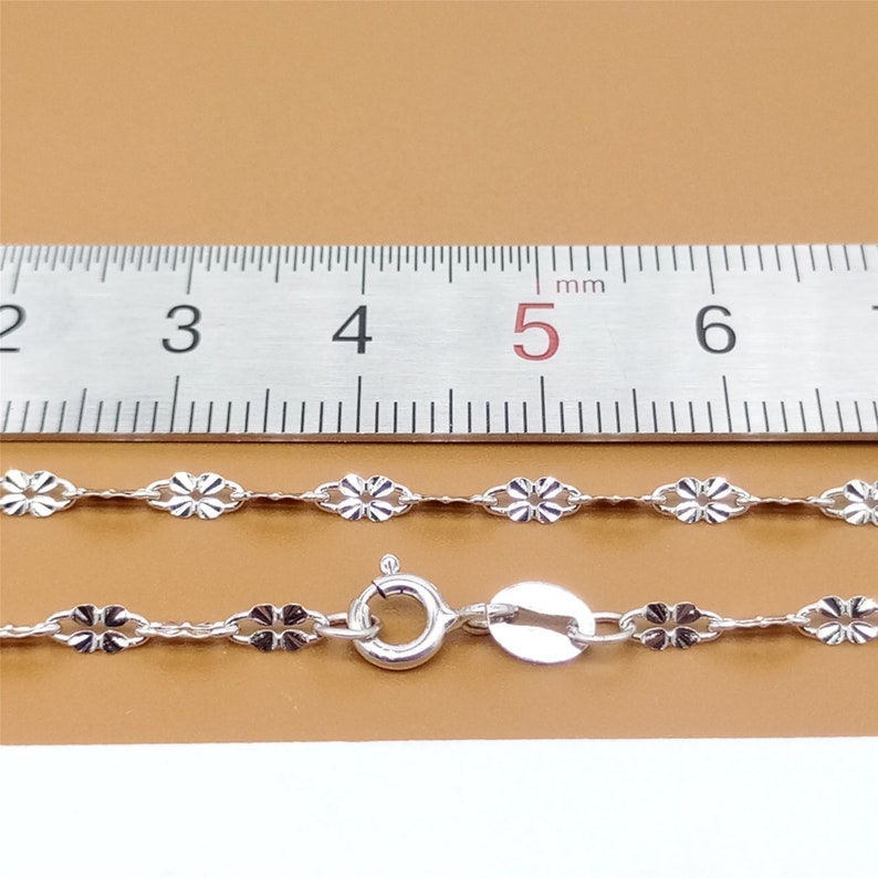 May include: A silver chain necklace with a delicate flower design. The chain is 5 mm wide and is measured against a ruler.