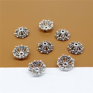 May include: Eight silver-toned metal bead caps with a decorative, intricate design. The caps are round and have a raised, circular pattern.