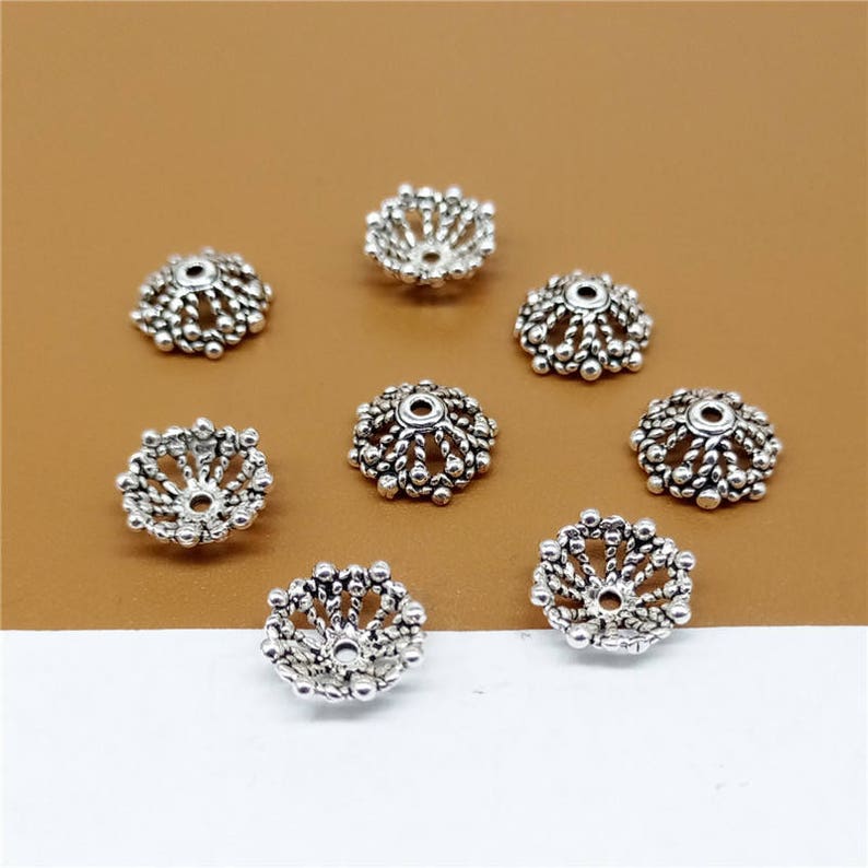 May include: Eight silver-toned metal bead caps with a decorative, intricate design. The caps are round and have a raised, circular pattern.