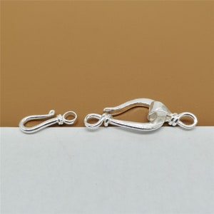 May include: A pair of silver-colored jewelry clasps. One is a hook and eye clasp, and the other is a larger, decorative clasp with a curved design. The clasps are shown against a brown and white background.