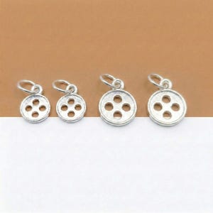 May include: Four small, silver-colored button charms with four holes each. The charms are round with a loop at the top for attaching to a chain or bracelet. The background is a mix of white and brown.