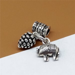 Könnte beinhalten: Ein silbernes Charm-Armband-Set. Das Set enthält einen Tannenzapfen, eine fassförmige Perle und einen kleinen Elefanten-Charm mit einem dekorativen Sattel. Die Charms sind durch eine kleine Kette verbunden.