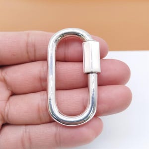 May include: A silver-colored, oval-shaped carabiner with a rectangular locking mechanism. The carabiner is held in a hand, showcasing its polished metal finish. This hardware item is suitable for various applications.