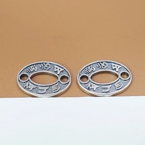 Puede incluir: Dos abalorios ovalados de plata con un diseño decorativo y dos agujeros. Los abalorios tienen caracteres japoneses.