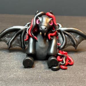May include: A small, sculpted dragon figurine with a matte black body, large spread wings, and vibrant red hair-like accents. The creature features piercing pink eyes, two small white fangs, and a curled tail with red detailing at the base.