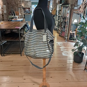 May include: A shoulder bag featuring a black and white houndstooth pattern on the main body, with solid grey fabric accents on the base and shoulder strap. The bag includes a long adjustable strap and a small white rectangular tag attached to the side.