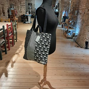 May include: A black and white tote bag featuring three vertical panels with different patterns: a polka dot section, a gingham check section, and a section with a repeating cat face print. The bag has two black shoulder straps and a small white tag.