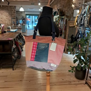 May include: A patchwork tote bag with a variety of fabrics, including denim, pink corduroy, and a pink and red plaid. The bag has navy blue straps and is displayed on a wooden mannequin.