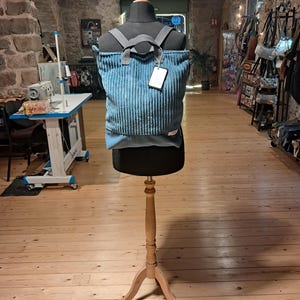 May include: A teal blue corduroy backpack with a rectangular shape, featuring a smooth solid blue base panel, two grey fabric shoulder straps, and a small white rectangular brand tag on the lower right corner, displayed on a black mannequin torso.
