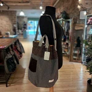 Puede incluir: Un bolso tote gris con una sección superior tejida marrón y un bolsillo marrón oscuro. El bolso tiene una correa gris para el hombro y una pequeña etiqueta adjunta. El bolso se exhibe en un maniquí en un entorno de tienda.
