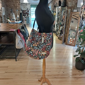 Könnte beinhalten: Eine Tragetasche mit floralem Muster, die in einem Geschäft auf einer Holzpuppe ausgestellt ist. Die Tasche hat einen dunklen Hintergrund mit einem Muster aus rosa, blauen und gelben Blumen. Das Geschäft hat einen Holzboden und Backsteinmauern, mit anderen Taschen und Artikeln.
