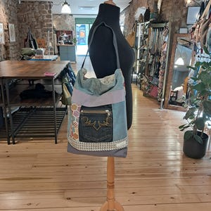 May include: A shoulder bag constructed from various fabric panels, including a dark denim pocket with gold stitching, a light blue textured section, a patterned hexagonal fabric strip, and a woven trim. The bag features a long shoulder strap.