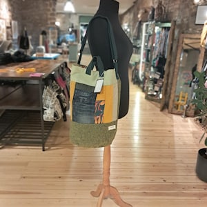 Könnte beinhalten: Patchwork-Tragetasche mit grünem, gelbem und Jeans-Design. Die Tasche hat einen Schultergurt und einen oberen Griff. Die Tasche wird auf einer Schaufensterpuppe in einem Geschäft präsentiert. Ein Etikett ist an der Tasche befestigt.