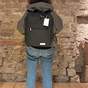 Könnte beinhalten: Eine Person trägt eine grüne Kapuzenjacke mit einem schwarzen Rucksack. Der Rucksack hat ein Reißverschlussfach und ein weißes Etikett. Die Person trägt eine blaue Jeans und hellblaue Turnschuhe.