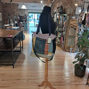 May include: A patchwork tote bag with a denim pocket and floral accents is displayed on a wooden mannequin. The bag features a light purple top, a black strap, and a tag. The background shows a store interior with wooden floors and stone walls.