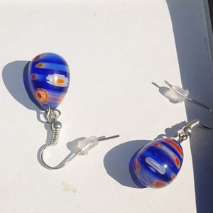May include: A pair of teardrop-shaped earrings with a blue, white, and orange pattern. The earrings have silver-colored hooks and clear plastic earring backs. The glass beads have a striped design with orange and white accents.