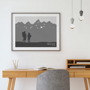 Puede incluir: Una impresi&oacute;n en escala de grises enmarcada que presenta una silueta de una cordillera y dos figuras. La impresi&oacute;n incluye el texto "Chris & Lizzie" y la fecha "12-1-16". La obra de arte se muestra sobre un escritorio de madera.