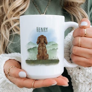 Peut inclure: Mug en céramique blanche avec une illustration à l'aquarelle d'un chien marron, de montagnes et le nom "HENRY". Le mug est tenu par une personne. Le design est imprimé sur le devant du mug.