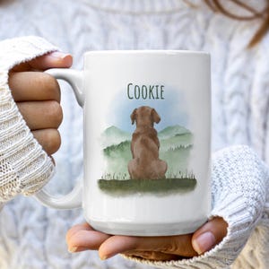 May include: White ceramic mug featuring a watercolor illustration of a brown dog sitting and looking at a mountain landscape. The name "COOKIE" is written above the dog in green letters.