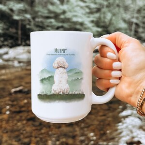 May include: White ceramic mug featuring a watercolor illustration of a dog with the name "Murphy" and the text "The Bestest Adventure Buddy." The dog is depicted sitting in front of a mountain scene. The mug has a large handle.