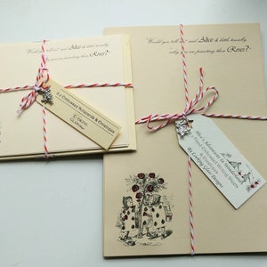 May include: Two sets of Alice in Wonderland themed stationery, each with a red and white twine bow and a charm. One set features a quote from the book, "Would you tell me, please, which way I ought to go from here?" and the other set features an illustration of Alice and the Queen of Hearts painting roses.