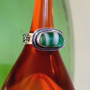 May include: A silver ring with a green and white oval-shaped glass stone set in a silver bezel. The ring band has a decorative cross design.