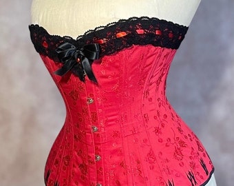 May include: A red satin corset with black lace trim and a black satin bow. The corset has a floral pattern and is designed with a pointed bottom.