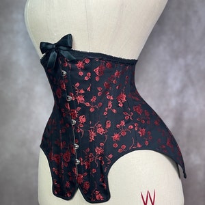 May include: A black and red corset with a floral pattern and a black bow at the top. The corset has a lace trim and is made of a satin-like fabric. The corset is on a white mannequin.