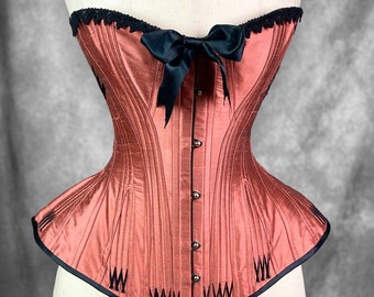 May include: A red satin corset with black lace trim and a black satin bow. The corset has a pointed bottom and is decorated with black stitching.