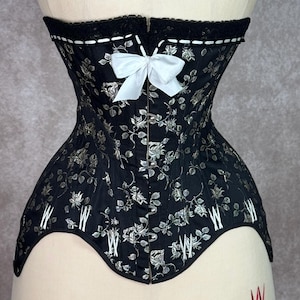 May include: A black corset with a white floral pattern and a white satin bow at the center front. The corset has a white lace trim at the top and a white stitching detail around the edges.