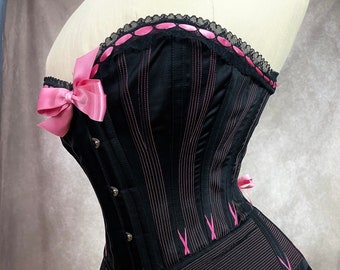 May include: A black corset with pink ribbon accents and a lace trim. The corset has a boned structure and a busk closure in the front.