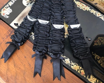 May include: Four black satin garters with silver metal accents and black ribbon bows. The garters are arranged on a wooden surface with a vintage sewing machine in the background.