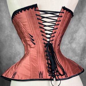 May include: A rust-colored corset with black lace trim and black lacing in the back. The corset has a boned structure and a pointed shape at the bottom.