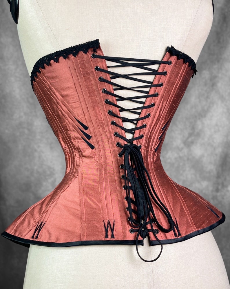 May include: A rust-colored corset with black lace trim and black lacing in the back. The corset has a boned structure and a pointed shape at the bottom.