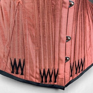 May include: A close-up of a red corset with black stitching and a black trim. The corset has a series of vertical lines and a decorative pattern of black "W"s on the bottom.