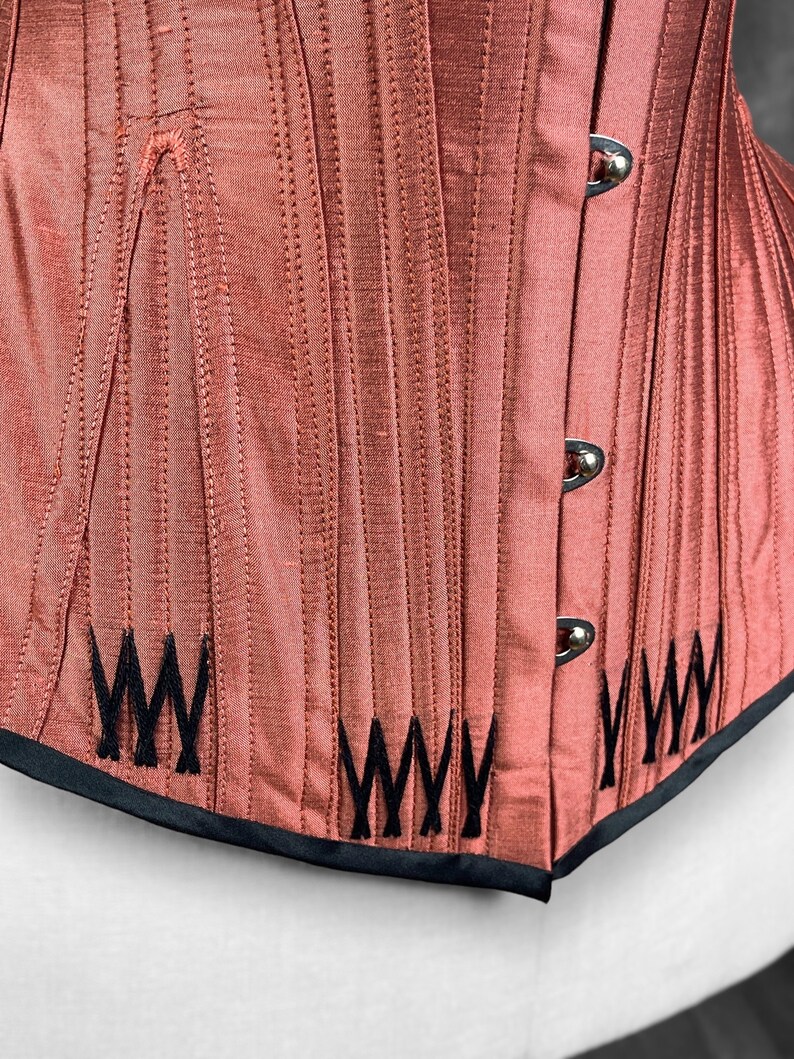May include: A close-up of a red corset with black stitching and a black trim. The corset has a series of vertical lines and a decorative pattern of black "W"s on the bottom.