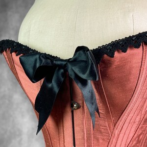 May include: A red satin corset with black satin ribbon lacing and black lace trim. The corset has a boned structure and a back closure.