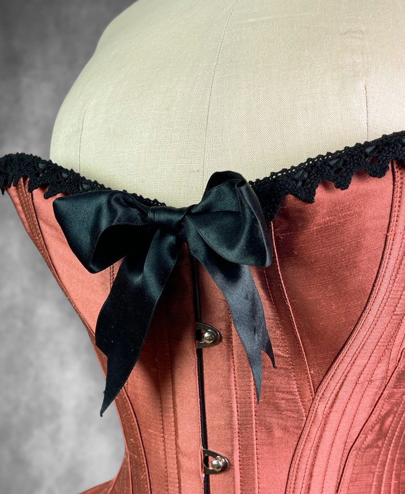 May include: A red satin corset with black satin ribbon lacing and black lace trim. The corset has a boned structure and a back closure.