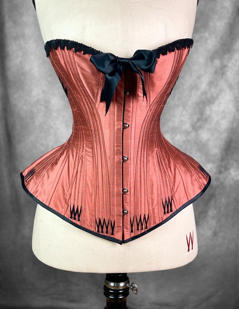 May include: A red satin corset with black lace trim and a black satin bow. The corset has a pointed bottom and is decorated with black stitching.