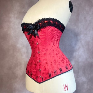 May include: A red corset with black lace trim and a black satin bow. The corset has a floral pattern and is on a white mannequin.