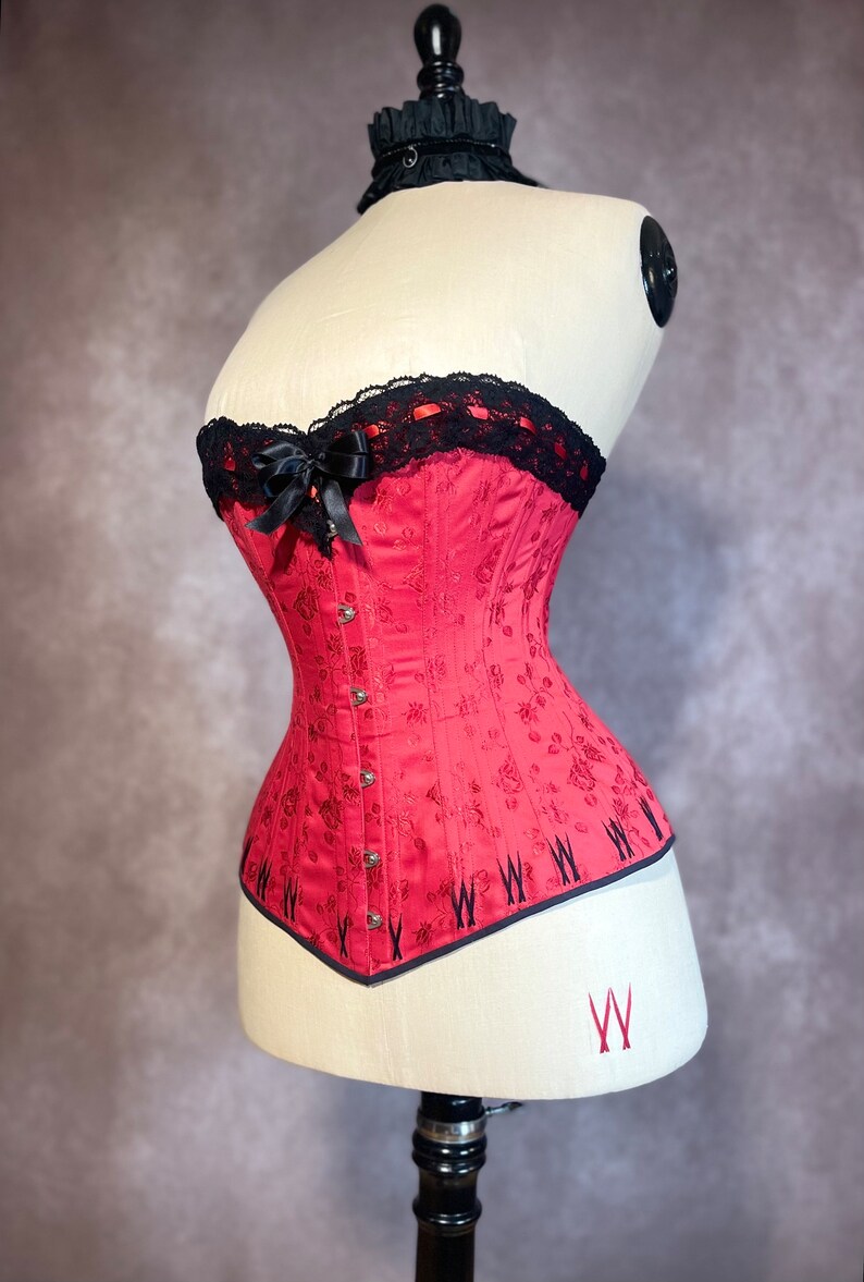 May include: A red corset with black lace trim and a black satin bow. The corset has a floral pattern and is on a white mannequin.