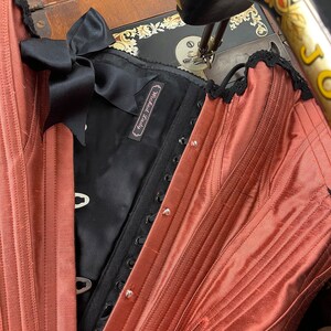 May include: A rust-colored satin corset with black lace trim and a black satin bow. The corset has a label that reads "Wicked Lovely".