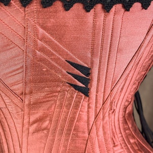 May include: A close-up of a red satin corset with black lace trim and black stitching. The corset has a decorative design on the front.