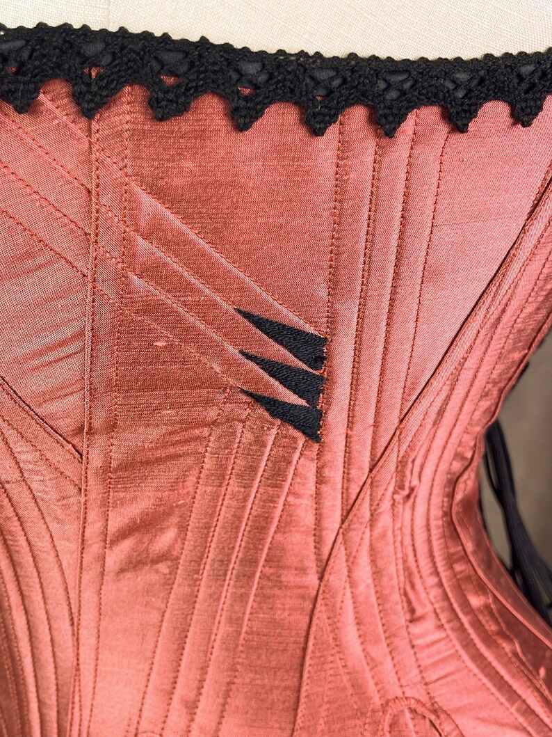 May include: A close-up of a red satin corset with black lace trim and black stitching. The corset has a decorative design on the front.